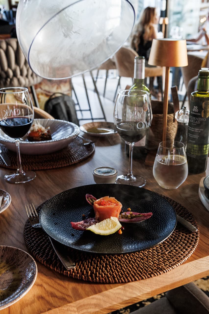 A plate of beef carpaccio at La Sala - thinly sliced cured beef under microgreens on a black ceramic dish, croquetas and olive-oil tasting plates around it, a glass of red wine at the table edge.