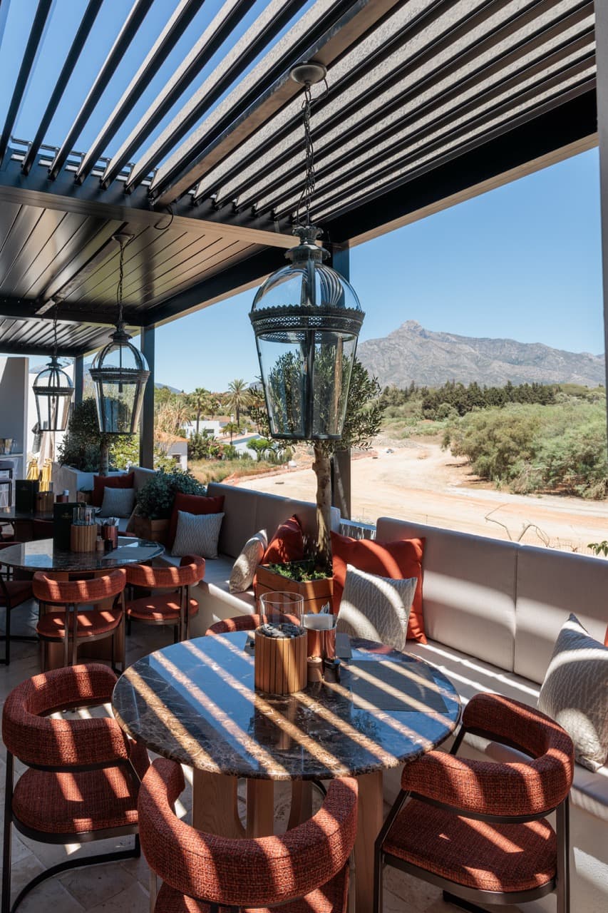 The VIP Terrace at La Sala - pergola roof and pendant lanterns over marble-topped round tables and burgundy chairs, La Concha mountain rising behind.