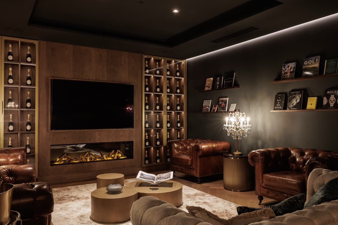 The Clubhouse Marbella — Chesterfield armchairs facing a wood-panelled hearth and screen, illuminated champagne wall on either side, low tungsten light.
