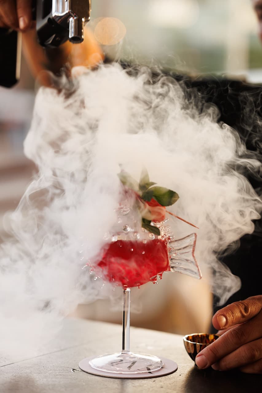 A signature cocktail at La Sala Puerto Banús - a long-stemmed fish-shaped coupe of red liqueur, a strawberry on the rim, dry-ice smoke from a torch above curling around the glass.
